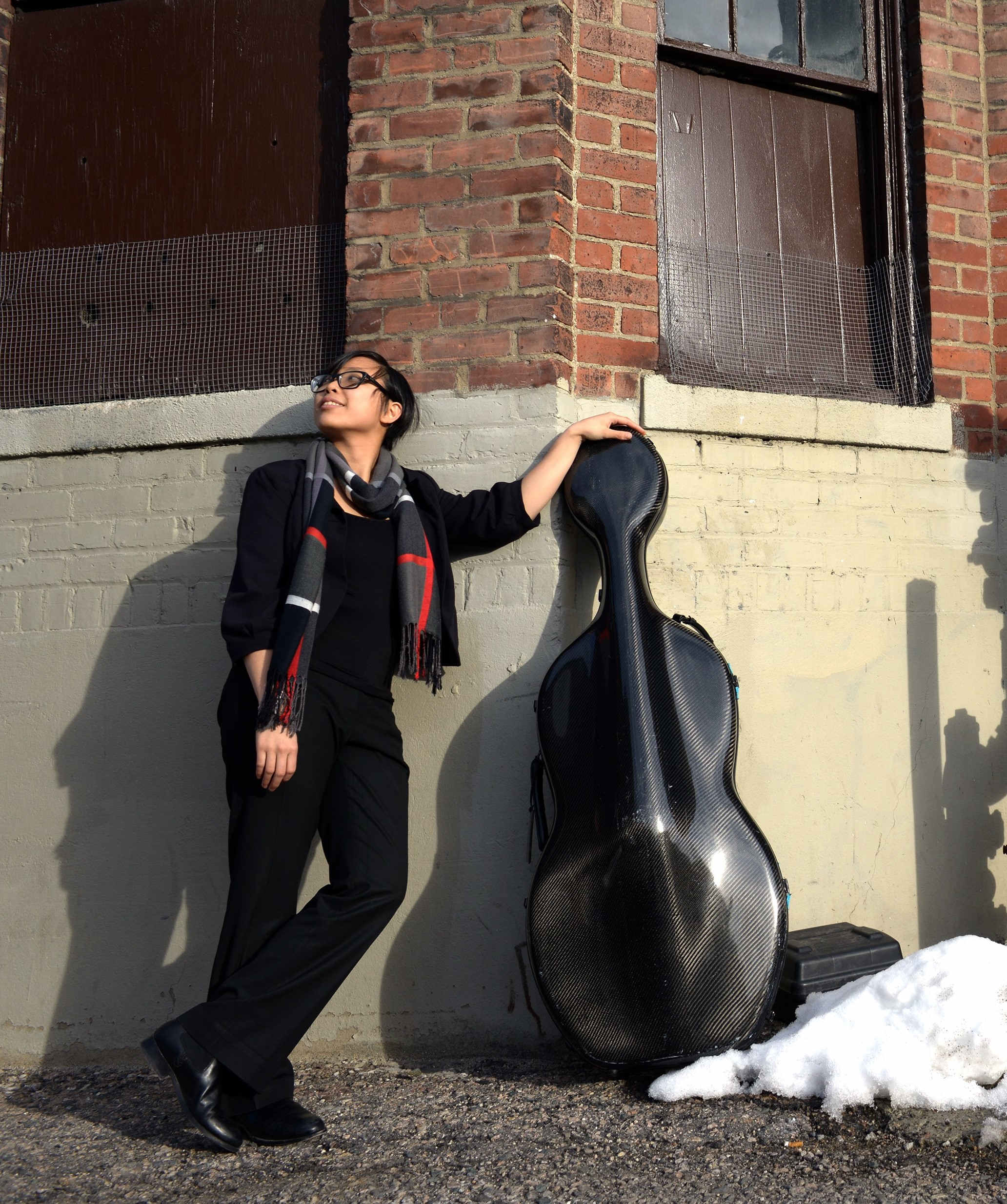 Sara leaning against building beside cello case